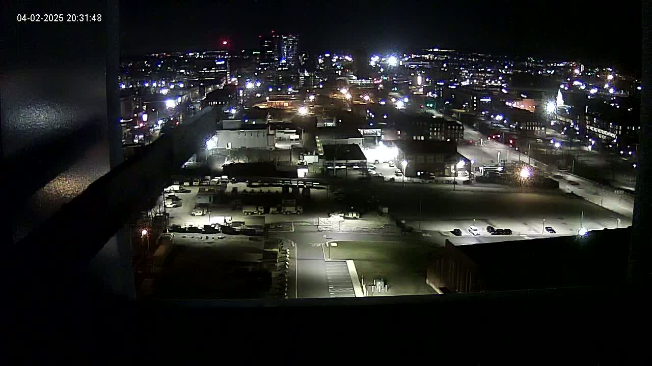 A nighttime, high-angle view of a city's industrial area, showing numerous buildings, parking lots with cars, and streetlights illuminating the scene under a clear, dark sky.
