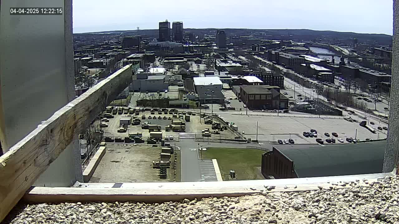 A high-angle, long shot on a sunny day shows a city landscape with numerous buildings, parked vehicles, and a large parking lot, viewed from a high vantage point that includes a portion of a building's roof and a wooden beam.