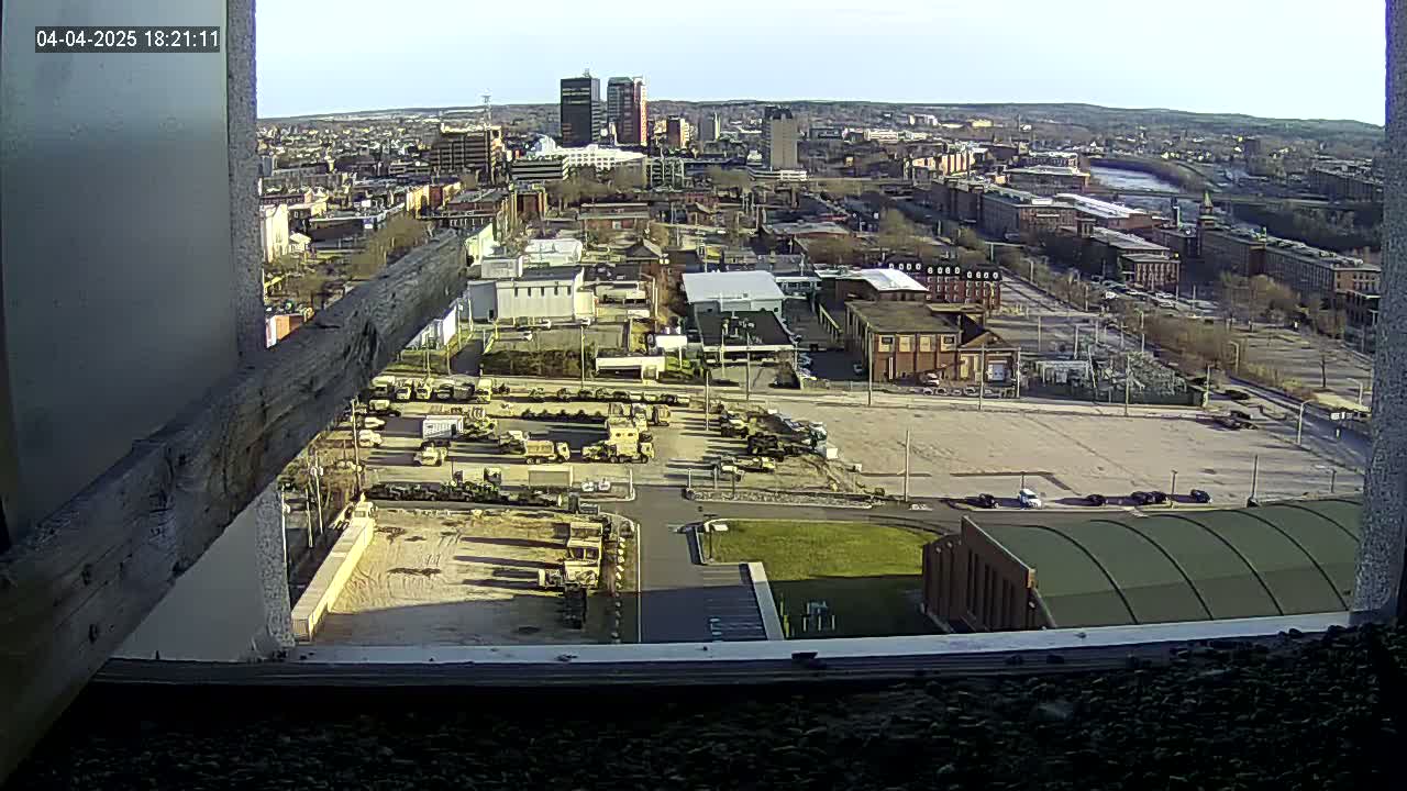 A high-angle, outdoor view on a sunny day shows a city with a large parking lot containing numerous military vehicles, and a river in the distance.