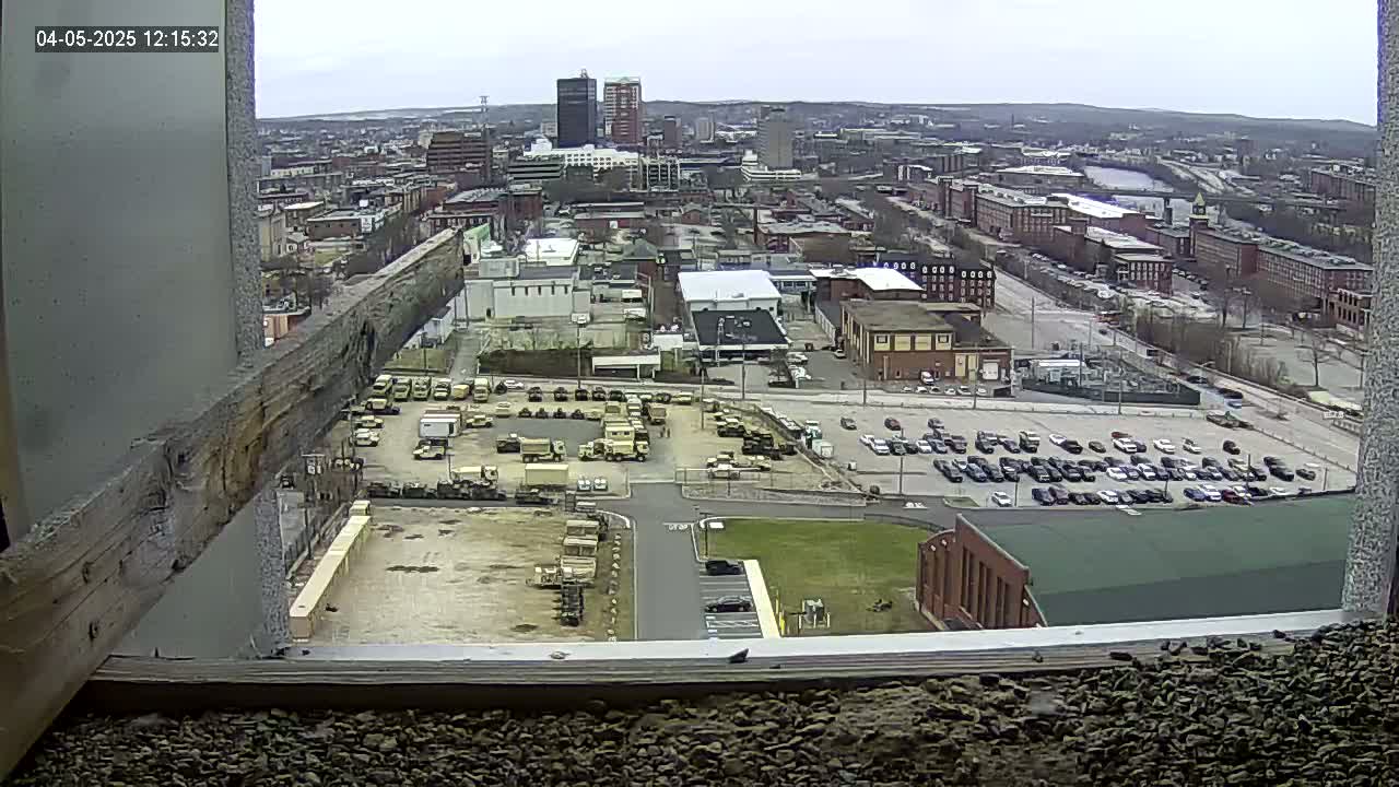A high-angle, long shot of a city on an overcast day, showing numerous parked vehicles, industrial buildings, and a large parking lot with many cars.