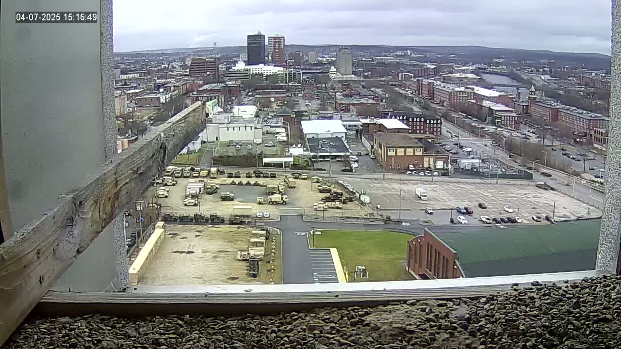 A high-angle, outdoor view on an overcast day shows a city with numerous buildings, a large parking lot containing several military vehicles, and a river in the distance.
