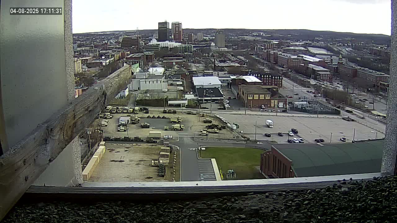 A high-angle view of a city on an overcast day, showing numerous buildings, parked vehicles, and a large empty lot, seen from a window.