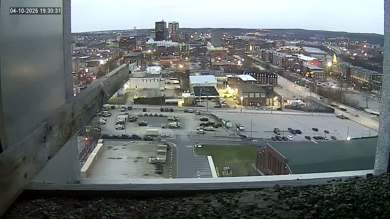 A high-angle, twilight view of a city with numerous buildings and vehicles in a parking lot, observed through a window with a weathered wooden beam in the foreground.
