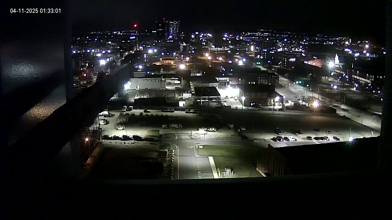 A nighttime, high-angle view of a city illuminated by streetlights and building lights, showing parking lots, buildings, and roadways.