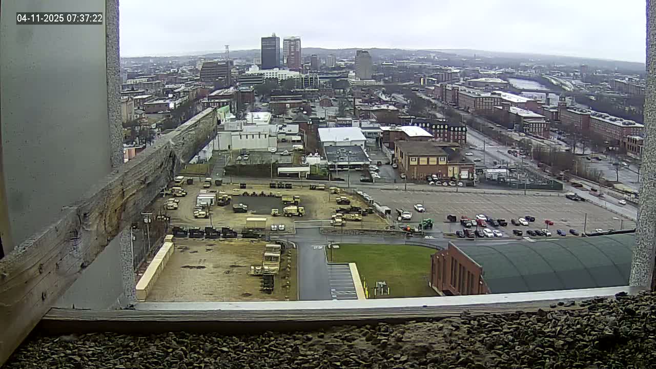 A high-angle, long shot on an overcast day shows a city with a large parking lot containing numerous vehicles, industrial buildings, and a scattering of military-style trucks in a nearby yard.
