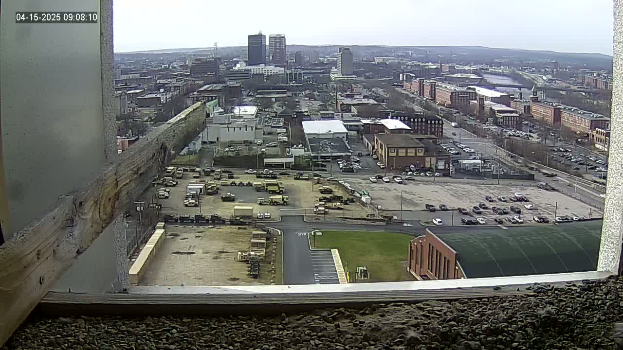 A high-angle, long shot of a city on an overcast day, showing a large parking lot filled with military vehicles and other industrial buildings, with a river visible in the distance.