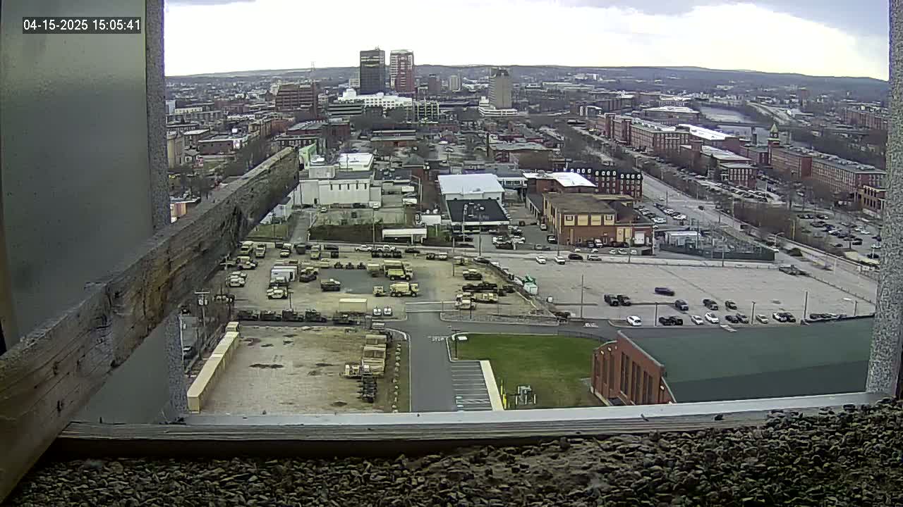 A high-angle, outdoor view on an overcast day shows a city with numerous buildings and parked vehicles, including a large lot filled with military-style trucks.