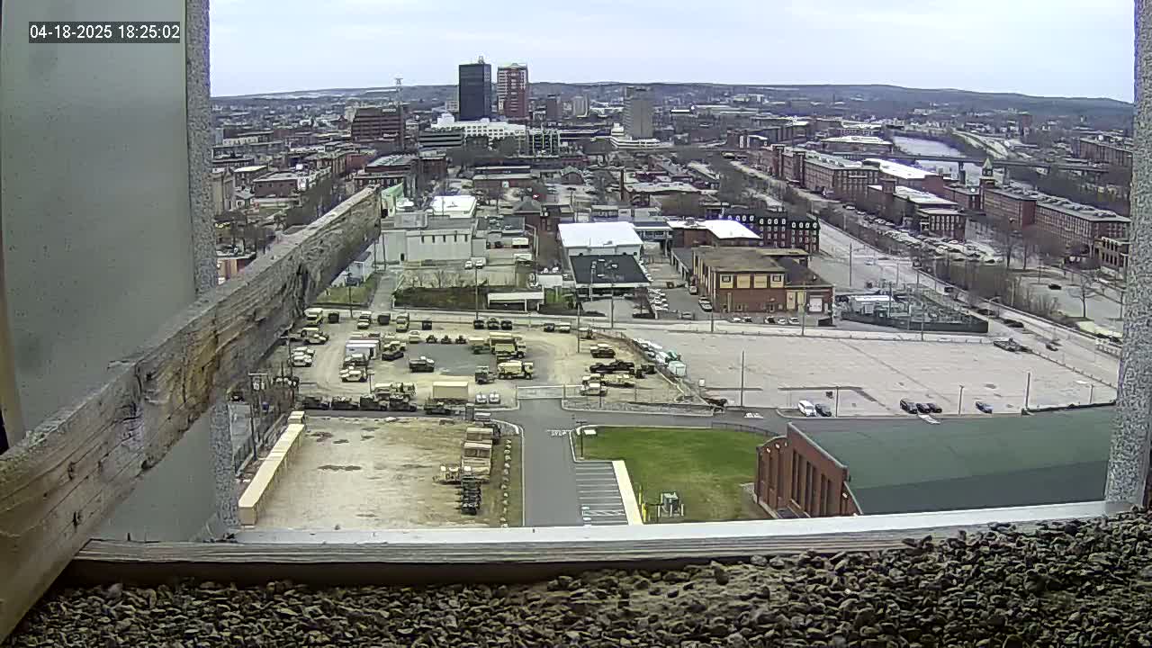 A high-angle, long shot on an overcast day shows a city landscape with numerous buildings, empty parking lots, and a collection of military vehicles parked in an organized lot.