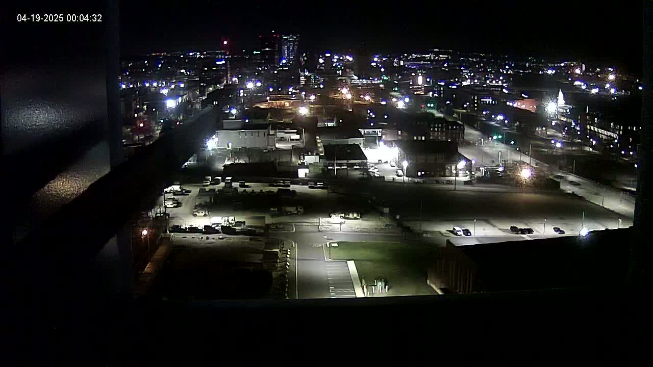 A nighttime, high-angle view of a city illuminated by streetlights and building lights, showing parking lots and buildings.