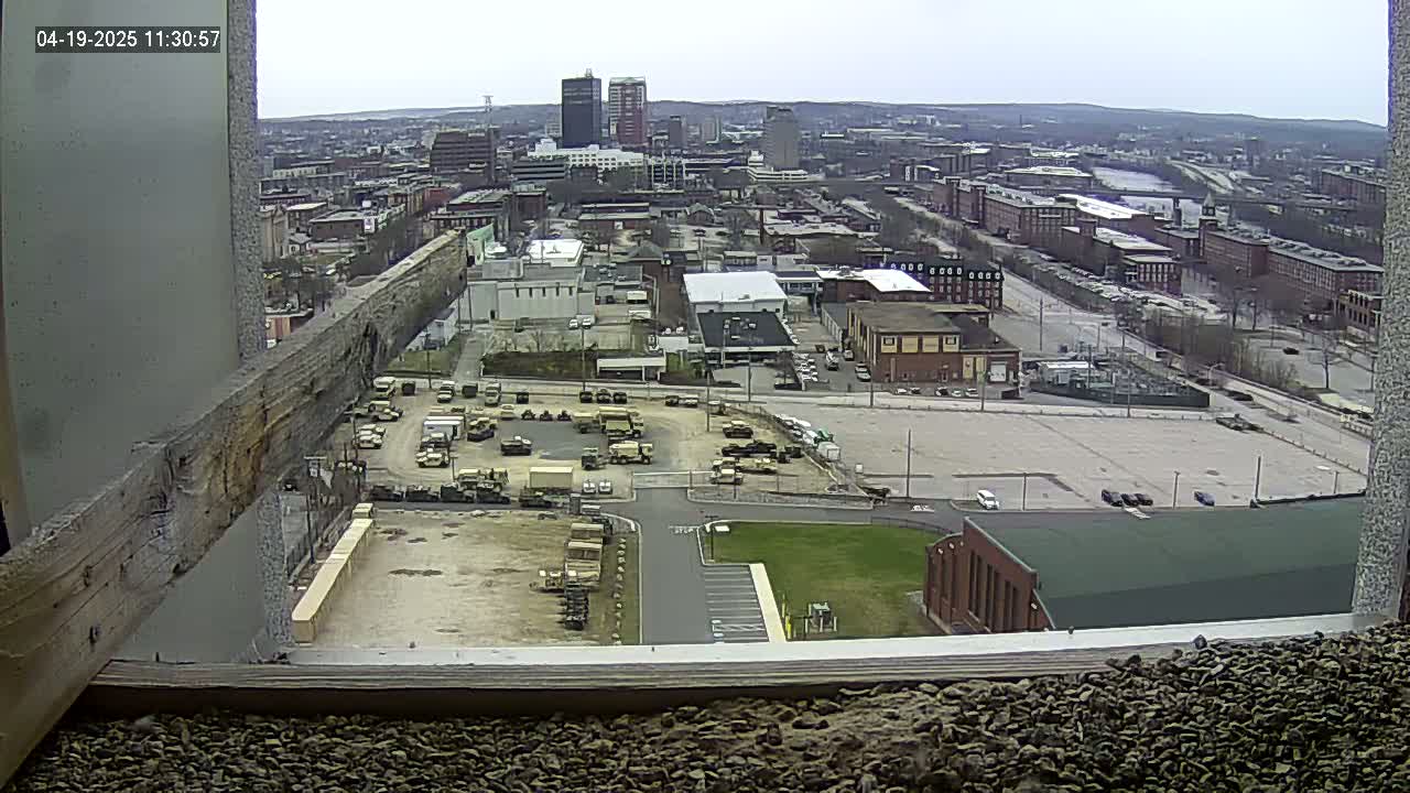 A high-angle, long shot on an overcast day shows a city with numerous buildings and a large parking lot filled with military vehicles.