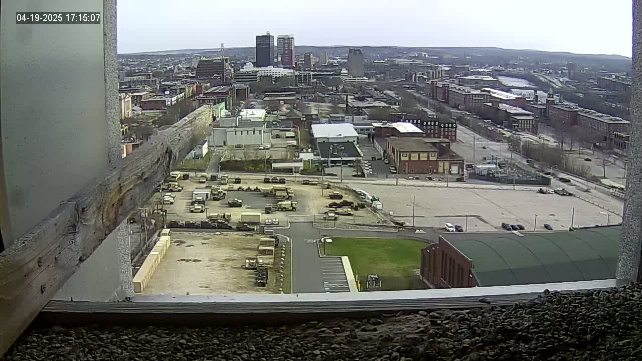 A high-angle, long shot on a partly cloudy day shows a city with military vehicles parked in a lot, and various buildings, including a large green-roofed building.