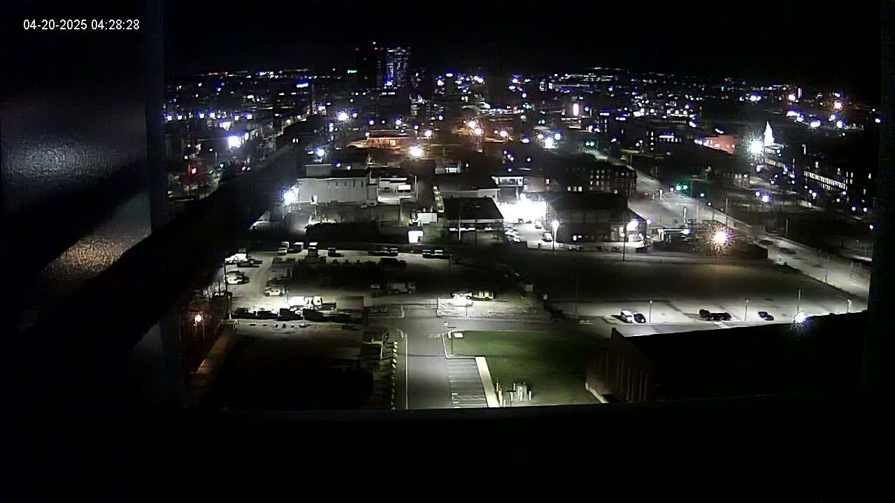 A nighttime, high-angle view of a city with numerous buildings and parking lots illuminated by artificial lights.