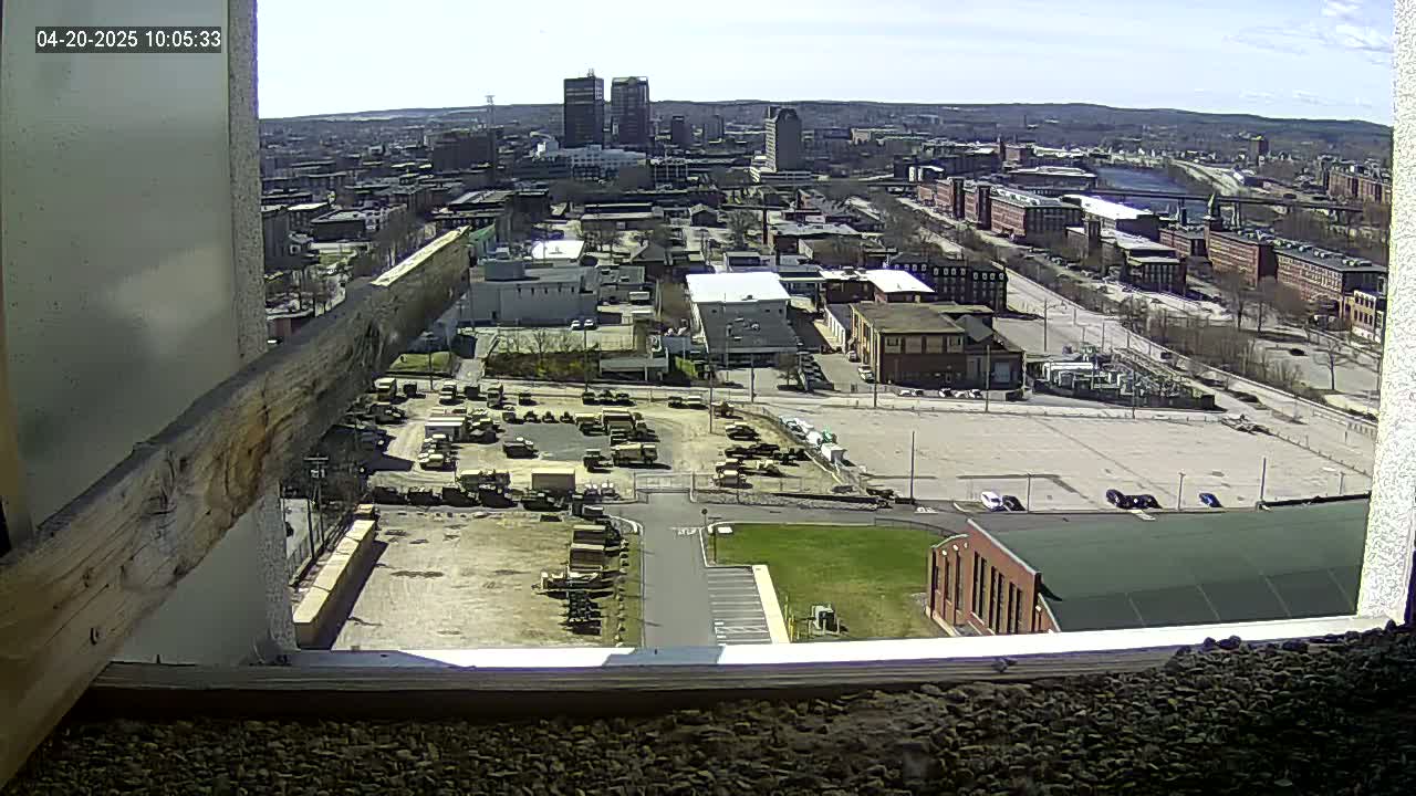 A high-angle, long shot on a sunny day shows a city with numerous buildings and a large parking lot containing military vehicles, viewed from a window.