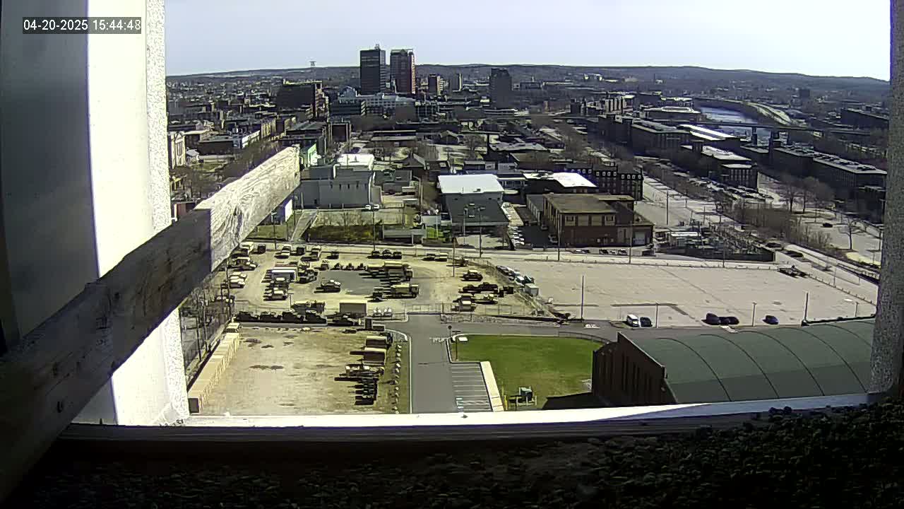 A high-angle, long shot on a sunny day shows a cityscape with a river, numerous buildings, and a large empty parking lot, viewed from a window with a wooden beam partially obscuring the view.