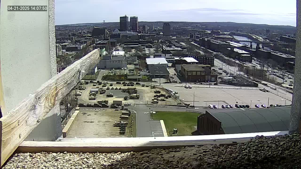 A high-angle, long shot on a sunny day shows a cityscape with numerous buildings, parking lots filled with vehicles, and a river in the distance.