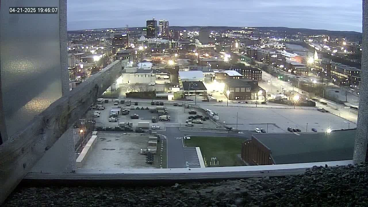 A twilight view from a high vantage point shows a city with numerous buildings and parking lots, with vehicles scattered throughout.