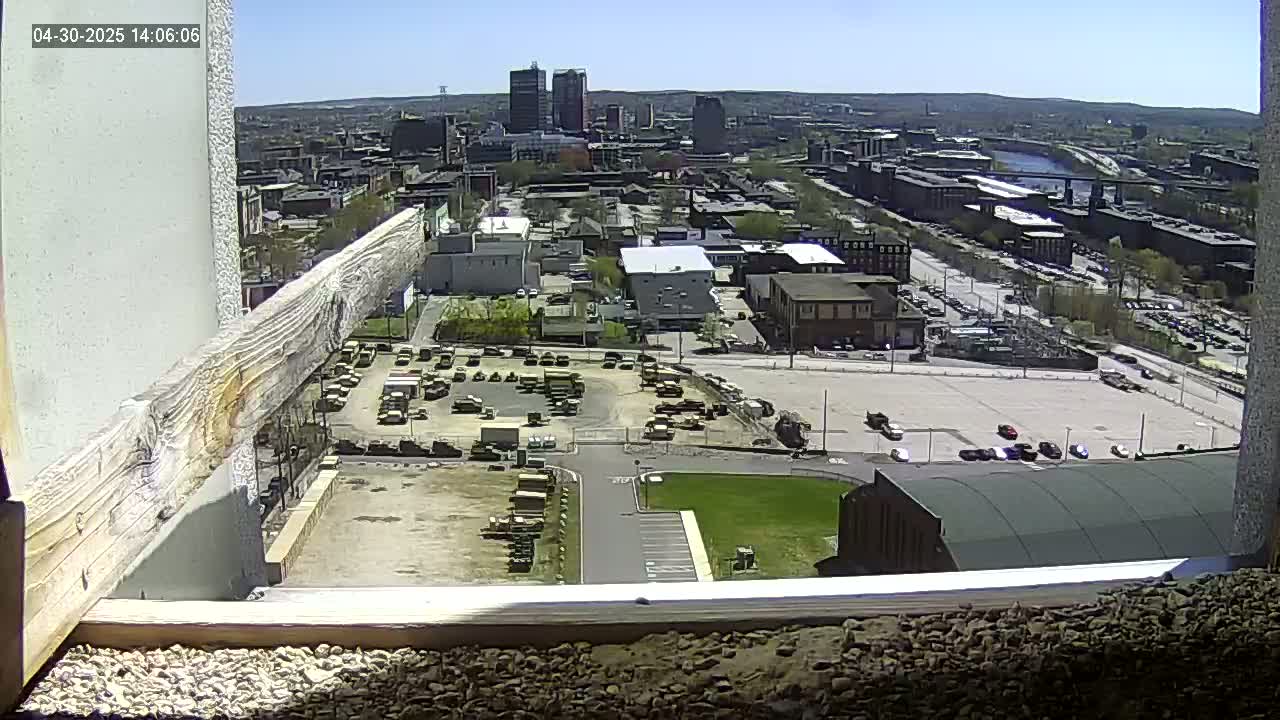 A high-angle, sunny view of a city with a large parking lot filled with numerous vehicles, including many military-style trucks, and a cityscape in the background.