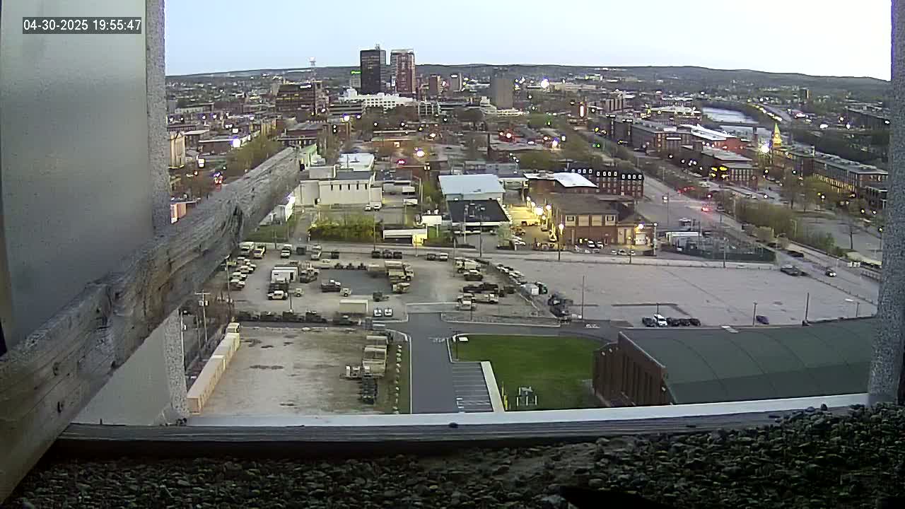 A high-angle, twilight view of a city with a large parking lot containing numerous military vehicles, overlooked by a building under construction.