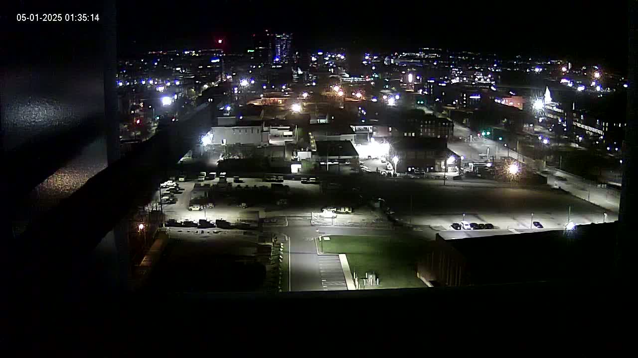A nighttime, high-angle view of a city landscape showing illuminated streets, buildings, and parking lots.
