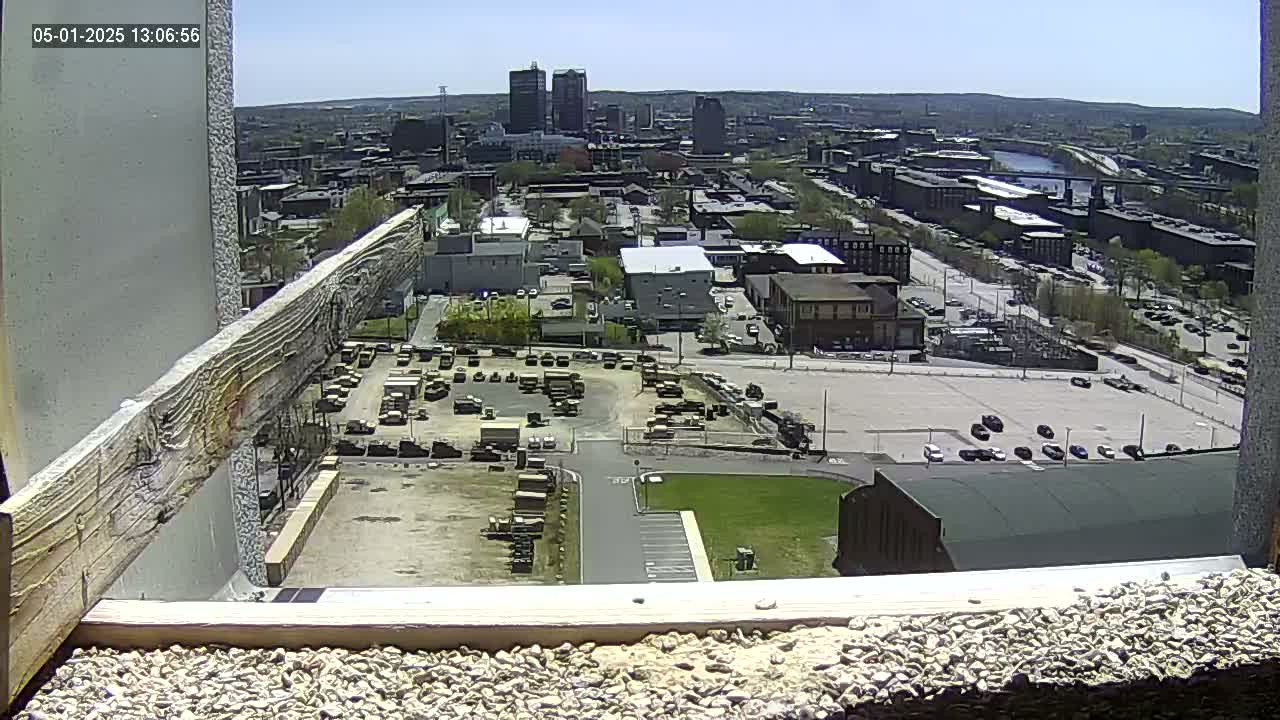 A high-angle, long shot on a sunny day shows a city with numerous buildings, parking lots containing vehicles, and a large expanse of empty space, all viewed from a high vantage point that includes a visible wooden beam in the foreground.