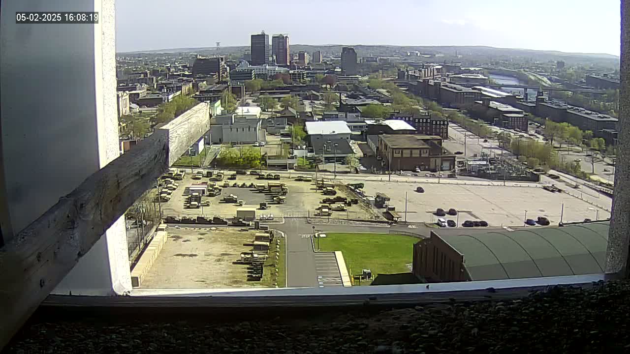 A high-angle, outdoor view on a sunny day shows a city with many buildings, a large parking lot filled with military vehicles, and a river in the distance.