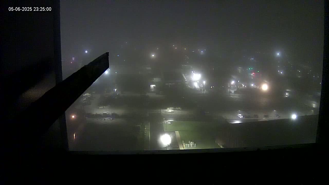 A foggy night scene is visible from a high vantage point, showing dimly lit buildings and vehicles in a parking lot.