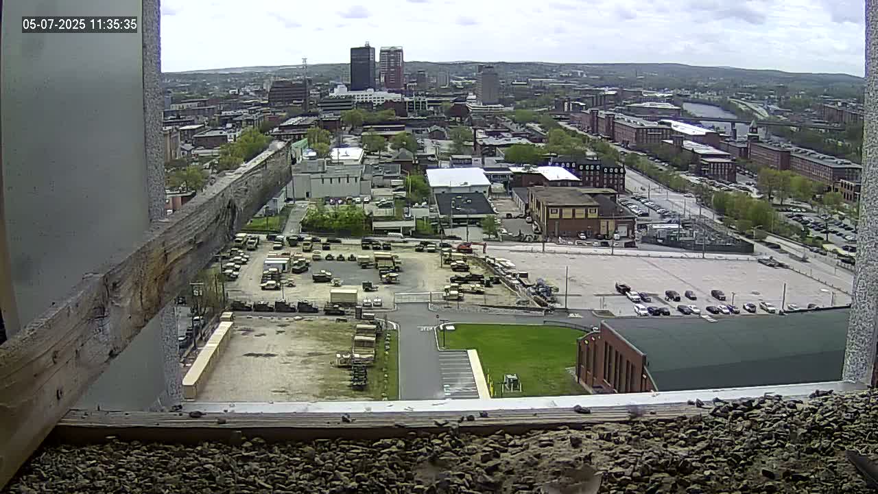 A high-angle, outdoor view on a partly cloudy day shows a city with numerous buildings, parked cars, and a large lot containing military vehicles.