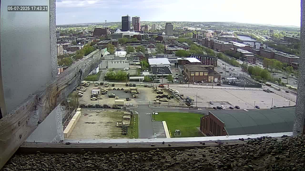 A high-angle, long shot on a partly cloudy day shows a city with military vehicles parked in a lot, surrounded by buildings and roads.
