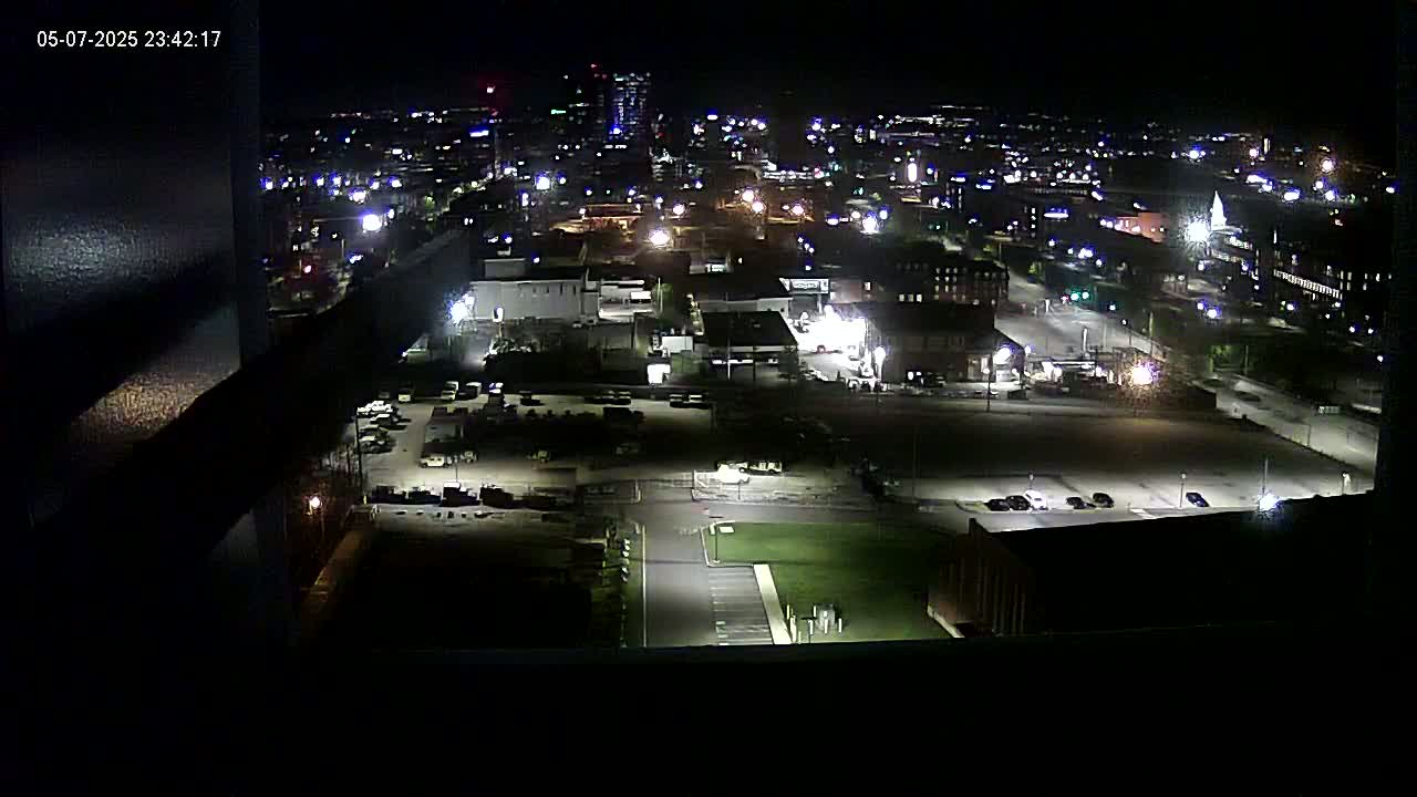A nighttime aerial view of a city's illuminated streets and buildings, including parking lots and a grassy area.