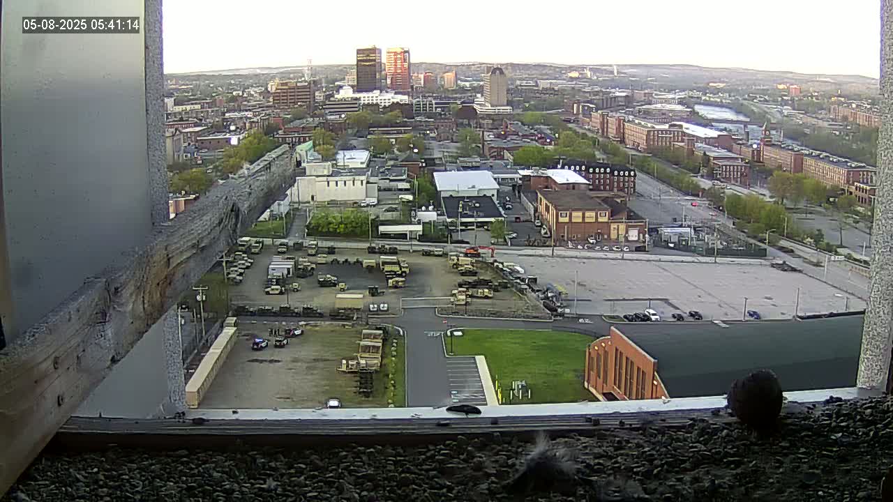 A high-angle, long shot of a city under a partly cloudy sky, showing a large parking lot filled with military vehicles, and a smaller lot with various other vehicles, all viewed from a window ledge.