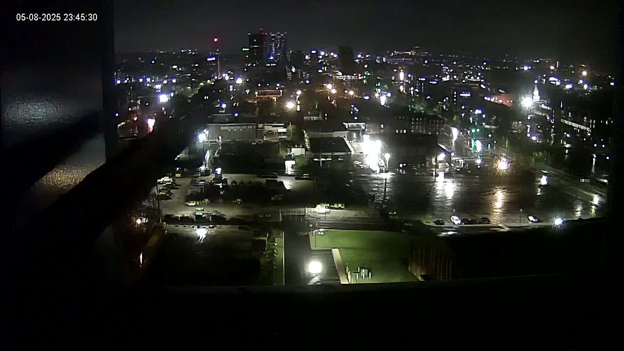 A nighttime view of a city with wet streets and a few cars visible under partly cloudy skies.