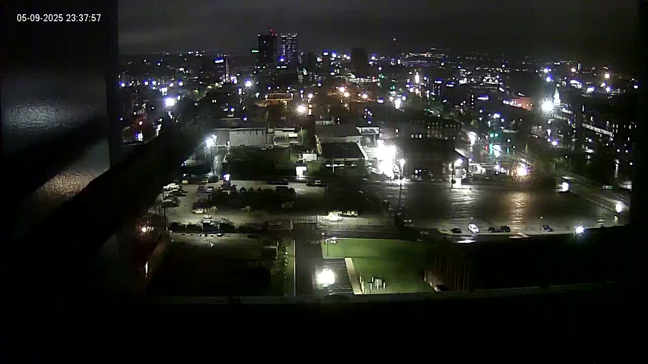 A nighttime, high-angle view of a city illuminated by streetlights and building lights, appearing wet from recent rain.