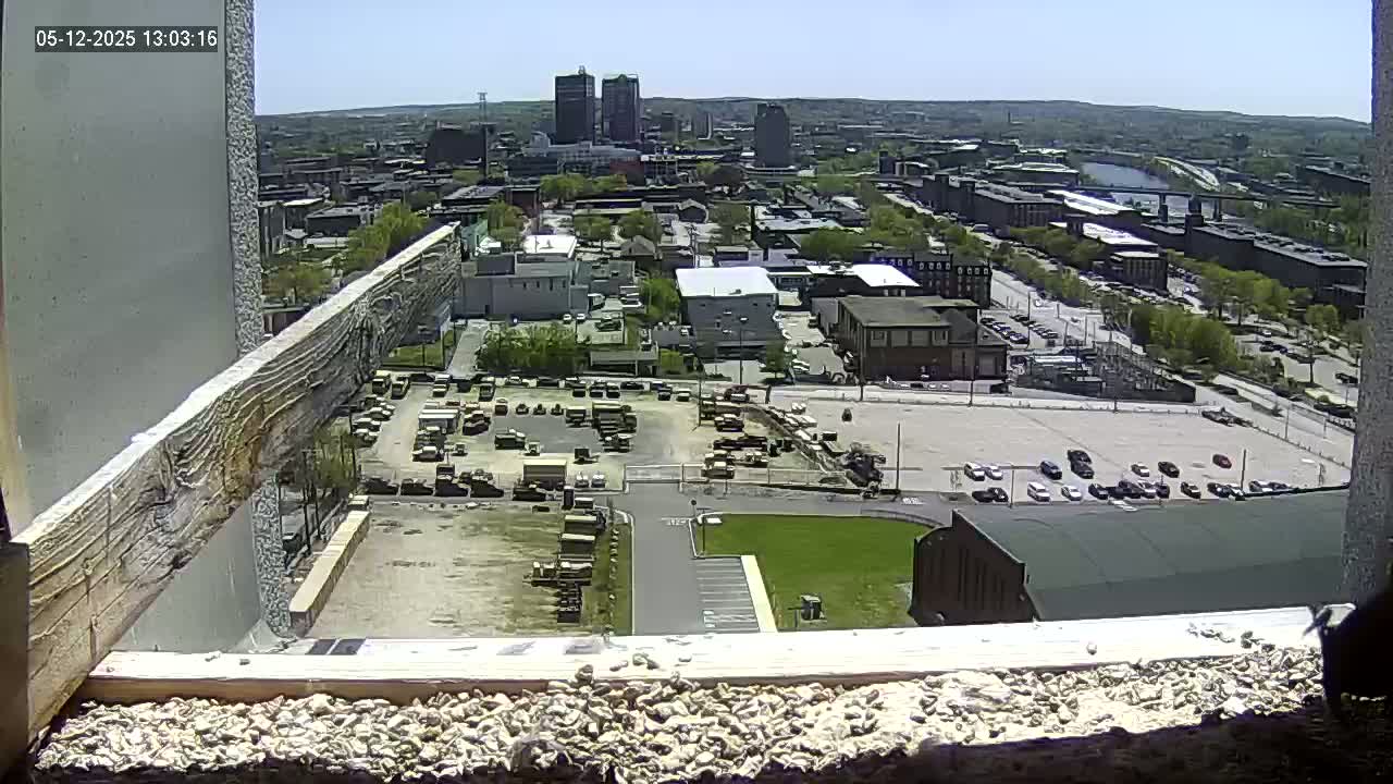 A high-angle, sunny view from a window shows a city with numerous buildings, parking lots filled with vehicles, and a large expanse of empty land, all under a clear sky.