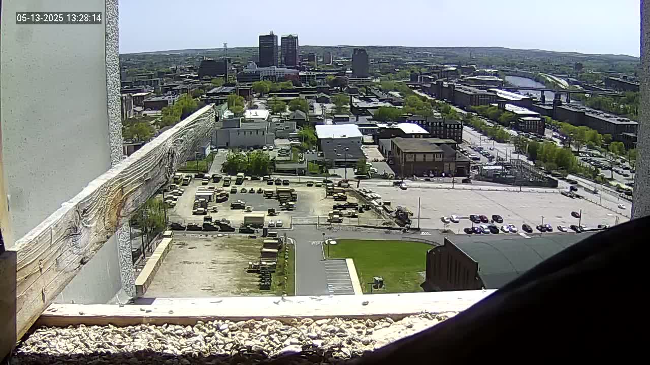 A high-angle, outdoor view on a sunny day shows a city landscape with numerous buildings, parking lots containing vehicles, and a large lot filled with what appears to be construction equipment or military vehicles.