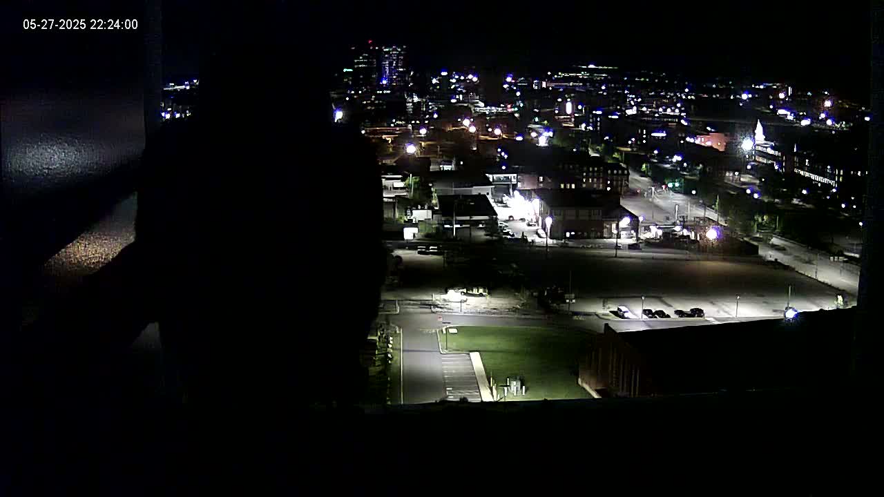 A nighttime, high-angle view of a city illuminated by streetlights and building lights, showing a mix of buildings, roads, and empty parking lots.