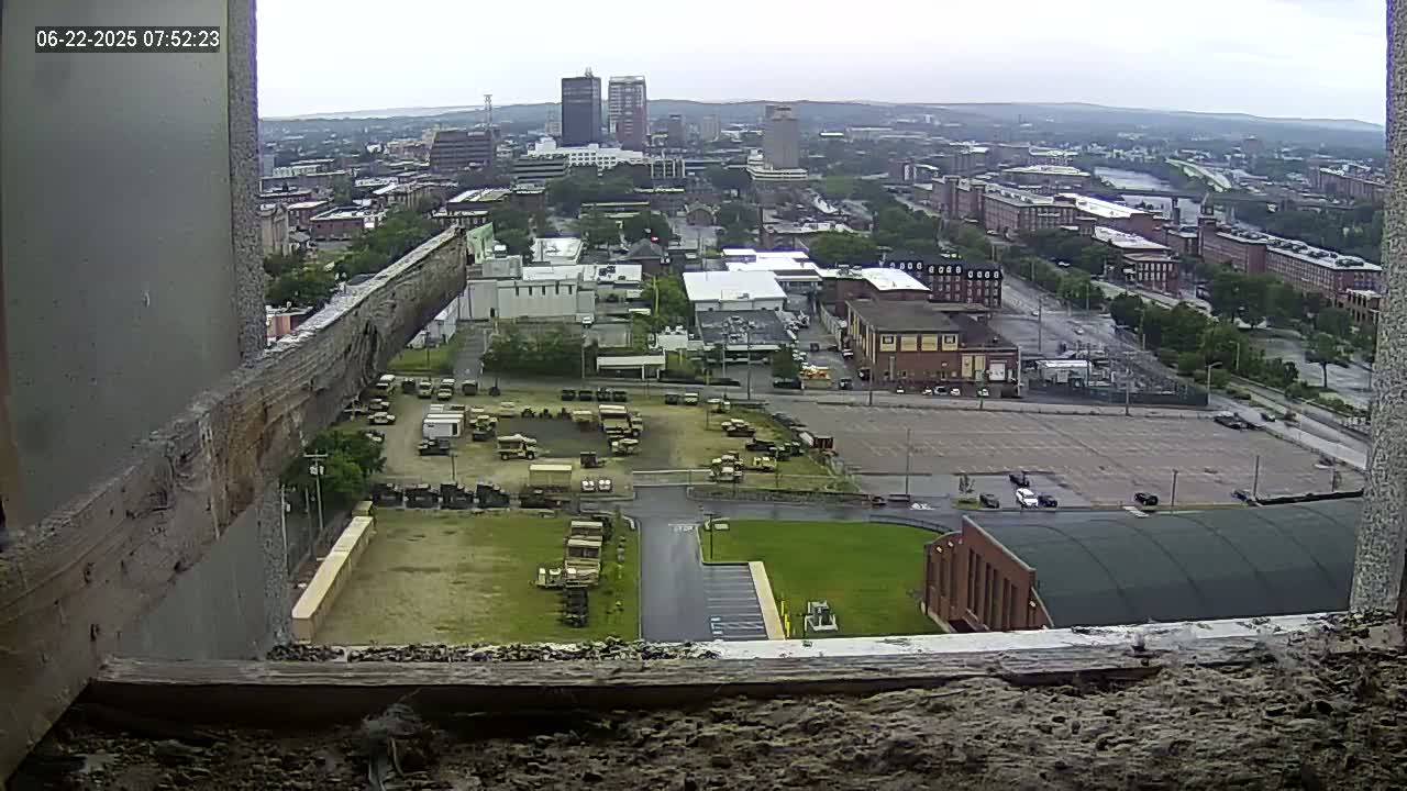 A high-angle view from a window shows a city under an overcast sky, with a large parking lot containing numerous military vehicles in the foreground.