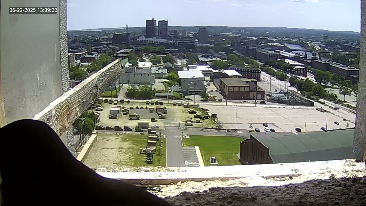 A high-angle view on a sunny day shows a city landscape with numerous buildings, a large parking lot containing military vehicles, and a smaller parking area next to a green-roofed building.