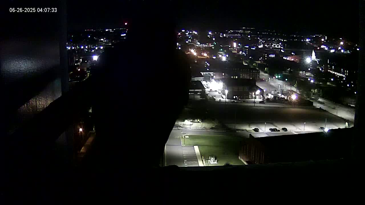 A nighttime, high-angle view shows a city illuminated by streetlights, with a dark, empty lot and a few parked cars in the foreground.