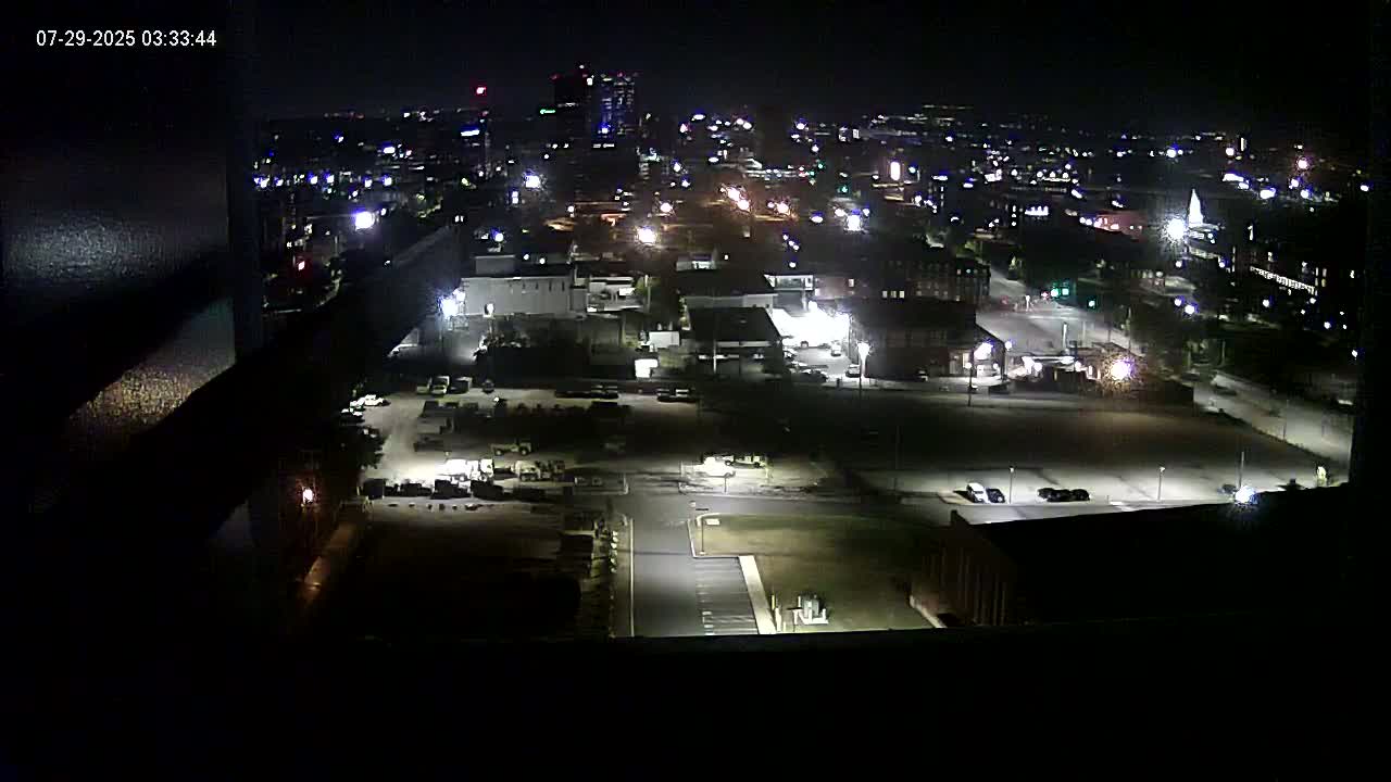A nighttime aerial view of a city, showing dimly lit streets, buildings, and parking lots.