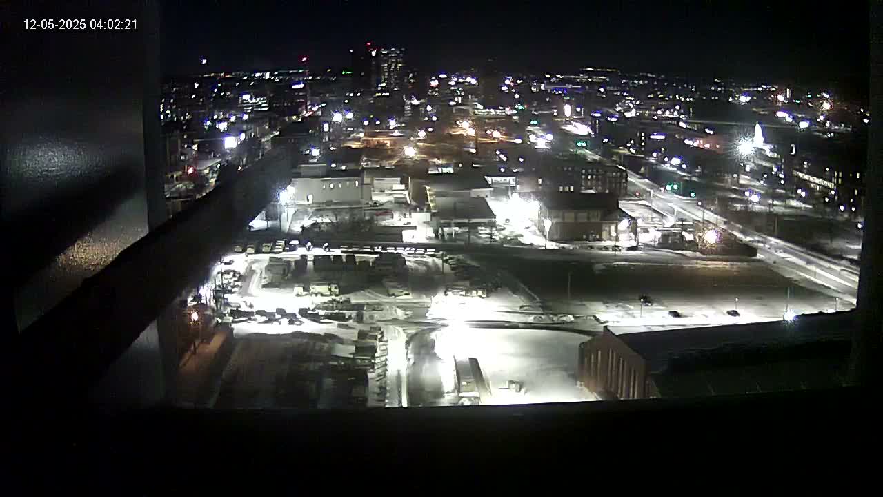 A clear nighttime wide shot from an elevated perspective showcases a brightly lit city with various buildings, active streets, and a large foreground area partially covered in snow.