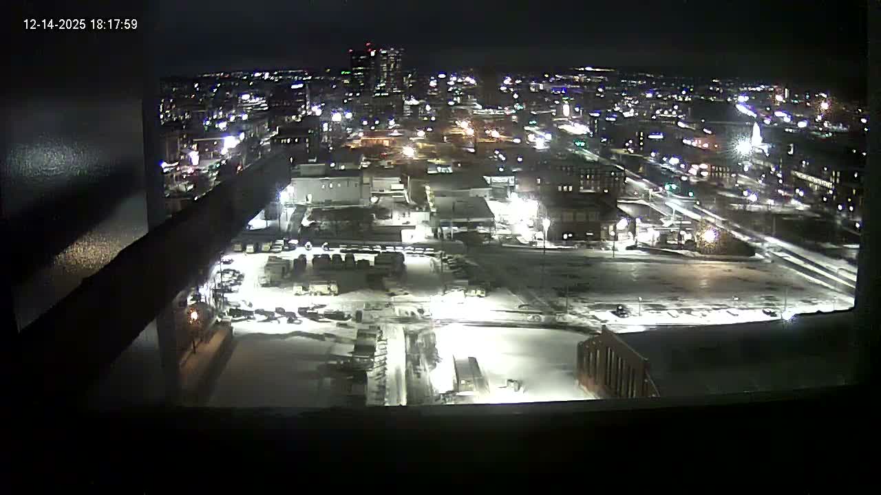 A clear nighttime wide shot from an elevated perspective showcases a brightly lit city with various buildings, active streets, and a large foreground area partially covered in snow.