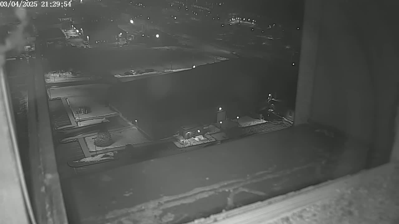 A nighttime, snow-covered view from a window shows a building complex with sparse lighting and some vehicles.