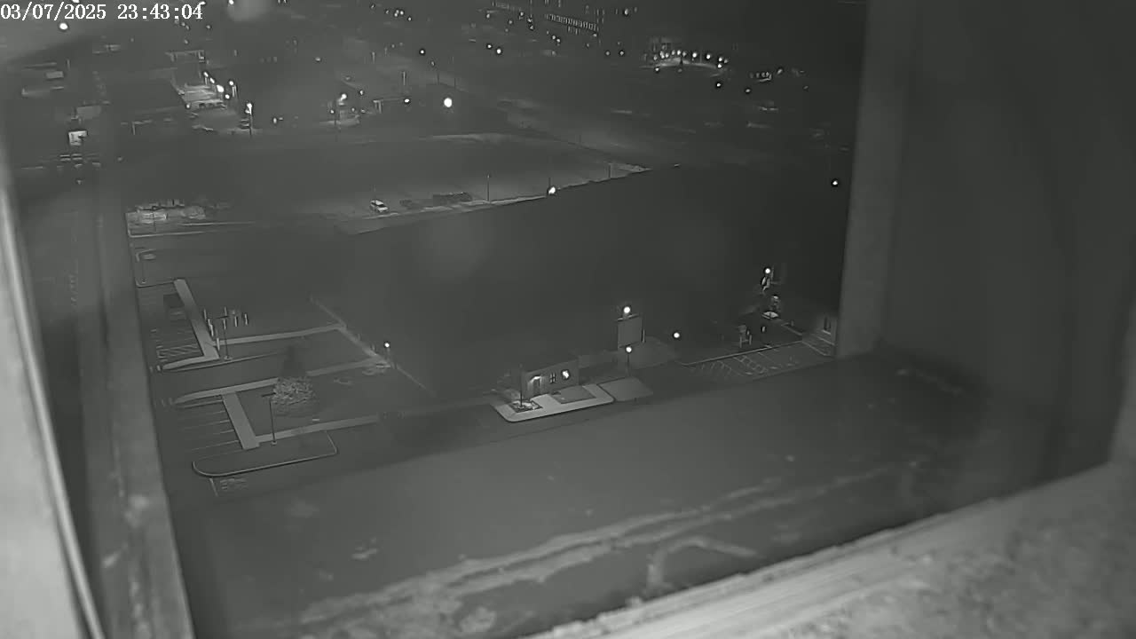 A nighttime, low-light view from a window shows a large building and surrounding parking lot in a mostly empty urban area.
