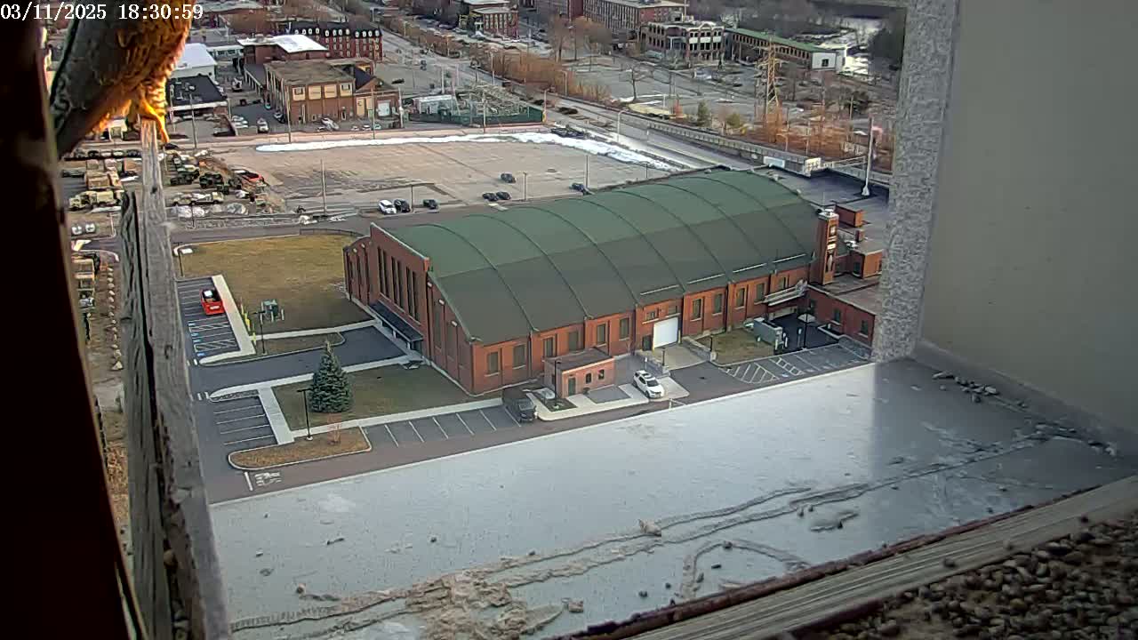A large brick building with a green arched roof is seen from above, in a mostly snow-free parking lot, with some snow patches remaining in the distance.