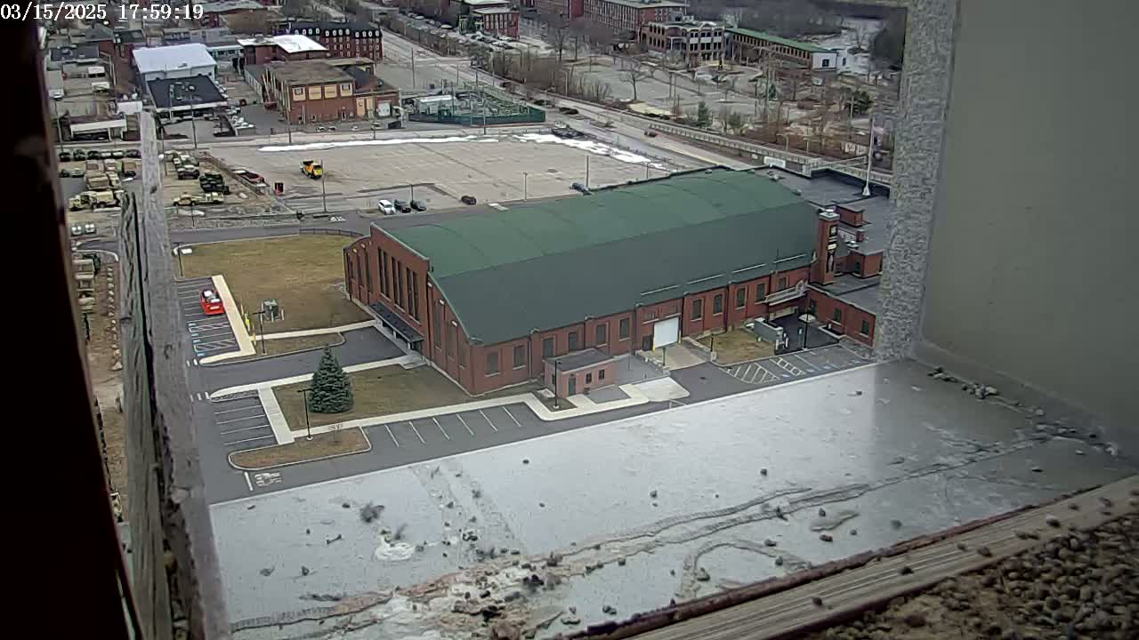 A large brick building with a green roof is seen from an elevated vantage point, with a mostly snow-covered parking lot and surrounding industrial area visible in the overcast day.