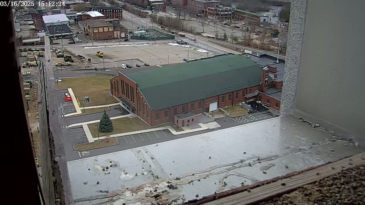 A high-angle view shows a large brick building with a green roof, situated in a parking lot next to other buildings, under a mostly clear sky.