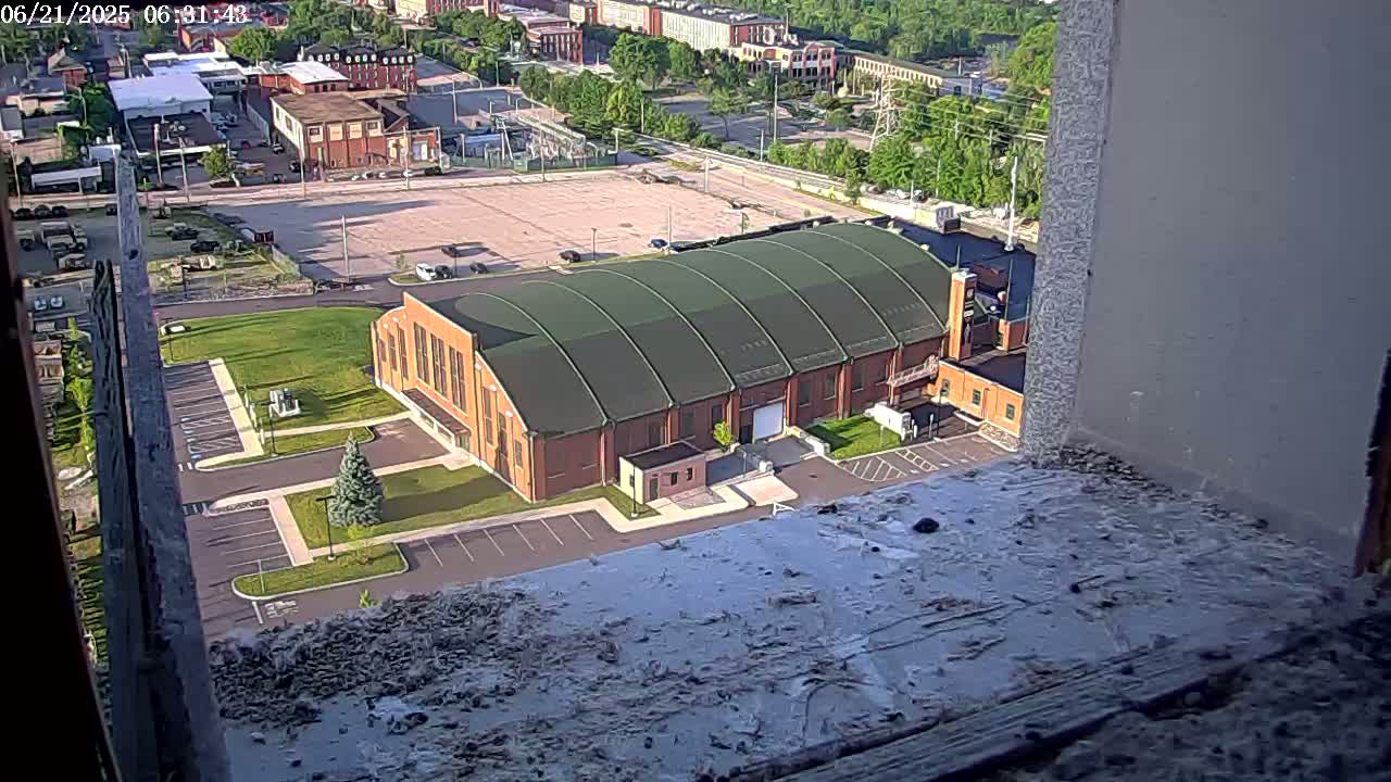 A high-angle view shows a large brick building with a curved green roof, surrounded by parking lots and some greenery, on a sunny day.