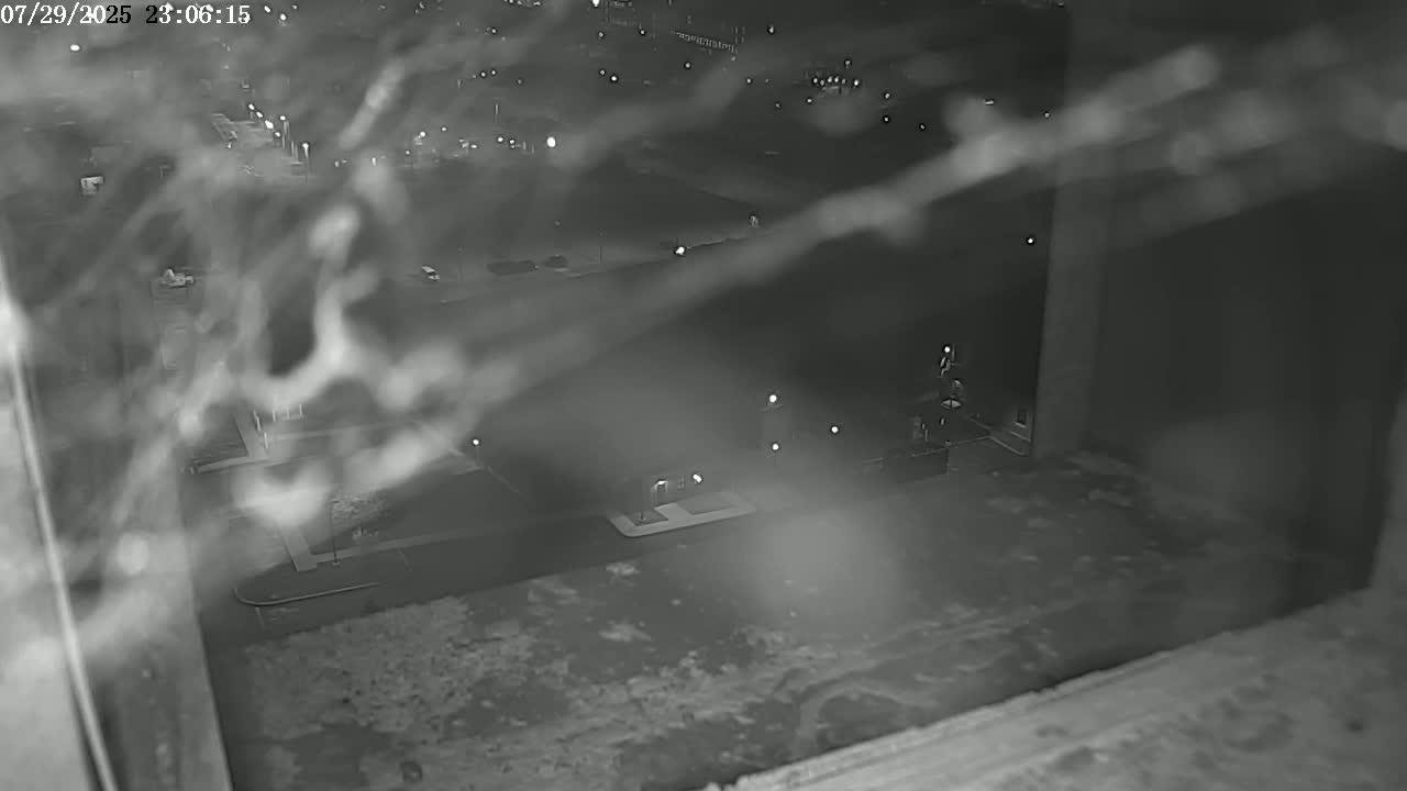 A nighttime view through a window shows a blurry, dimly lit street scene with buildings and parked cars obscured by a web-like pattern.