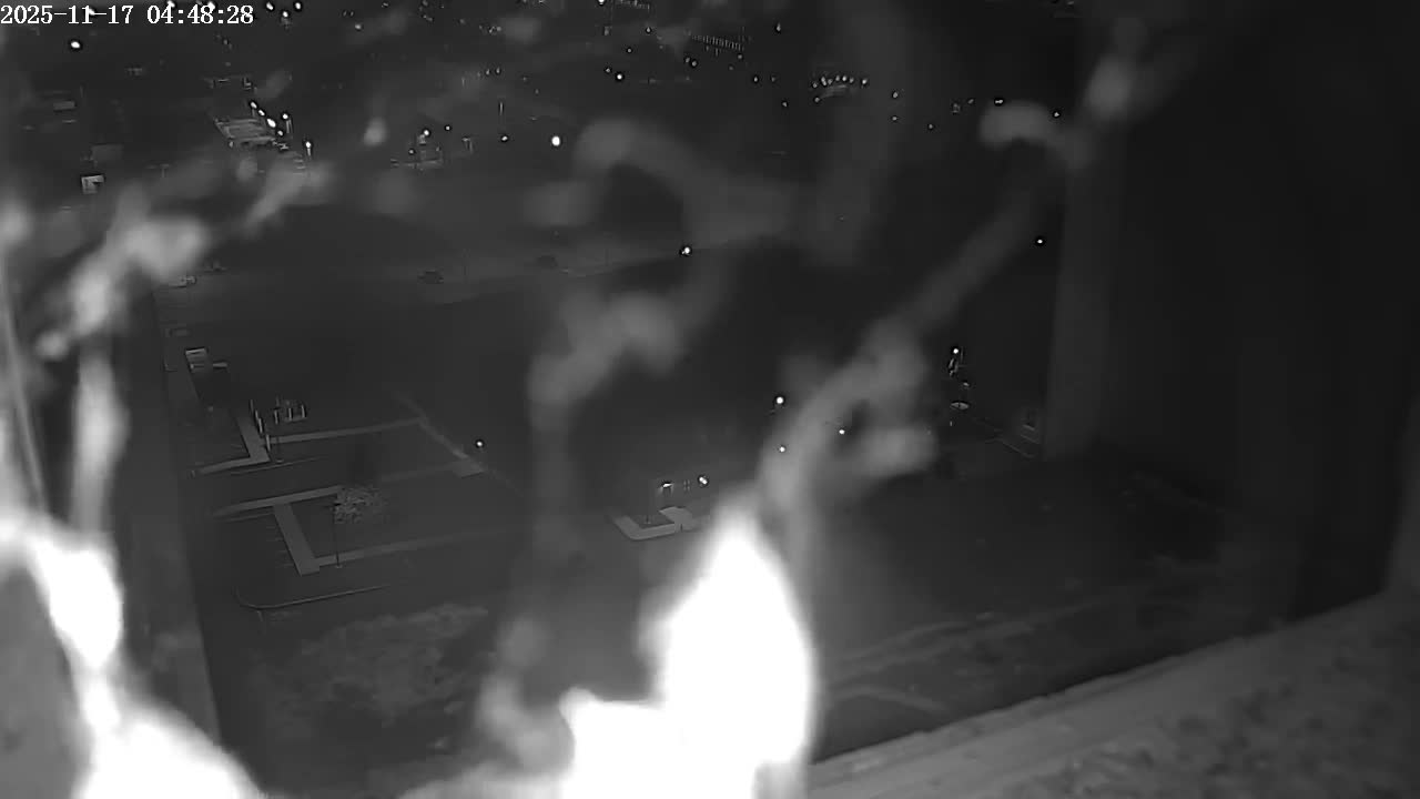 An elevated, grayscale nighttime view of a city street and buildings is partially obscured by blurry raindrops on the camera lens, indicating a rainy evening.