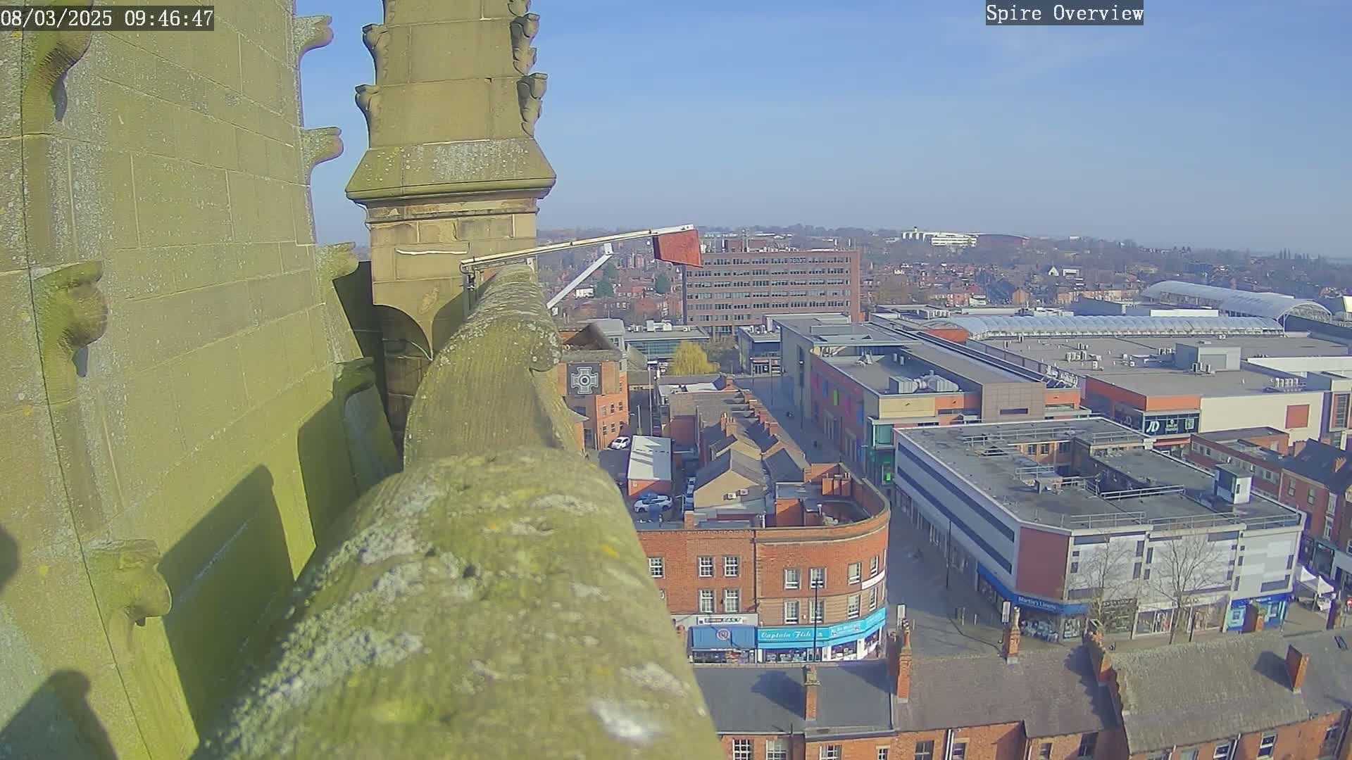 A high-angle view from a stone spire on a sunny day shows a city center with buildings and a shopping mall.